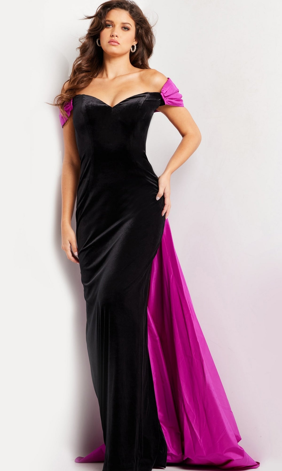 A woman stands against a plain white background wearing the Long Prom Dress 37375 by Jovani, featuring an off-the-shoulder black velvet fitted silhouette with a striking magenta train.